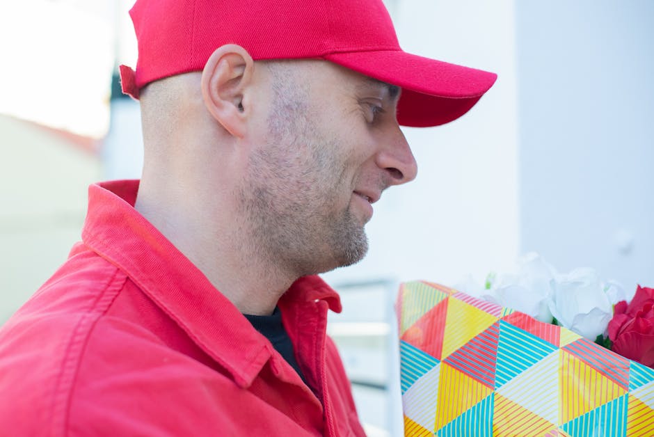 A delivery person in a red cap and matching red jacket is holding a colorful, geometric-patterned gift bag filled with white and red flowers. The person is smiling slightly and appears to be engaged i