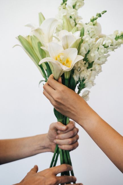 A person holding a bouquet of fresh white flowers, including lilies and delphiniums, arranged with green stems and leaves. The bouquet is wrapped in transparent tissue paper, with a clean, minimal pre