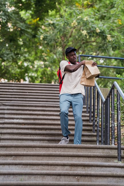 A young man with dark skin, wearing a black cap, white T-shirt, light blue trousers, and grey shoes, is descending outdoor concrete stairs in a park-like setting surrounded by lush green trees. He is