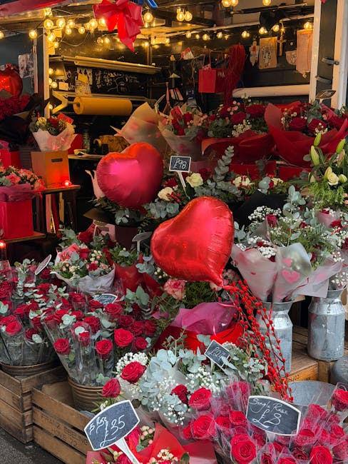 A vibrant flower display featuring a variety of red and white blooms, including roses and smaller filler flowers, arranged in wicker baskets and wrapped in white and red paper. The arrangement is acce