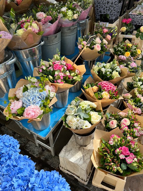 A display of freshly arranged floral bouquets on a blue table, featuring a variety of pastel-colored flowers including pink roses, white lisianthus, light blue hydrangeas, and yellow accents, wrapped 
