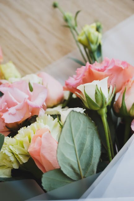 A close-up view of a floral bouquet featuring soft pink roses, cream-colored lisianthus, and green foliage, arranged in a natural style with some flowers partially open revealing layered petals. The b