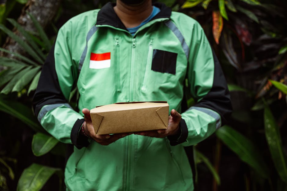 A delivery person wearing a green and black jacket with a red and white patch on the chest, holding a rectangular brown paper parcel with folded edges, standing amidst lush green foliage. The jacket f