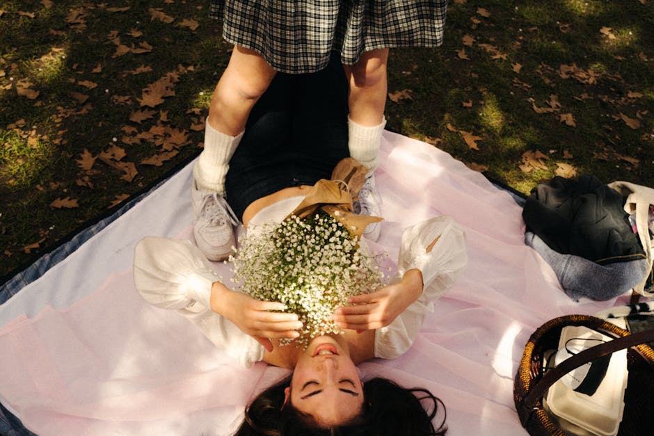 A young woman with long dark hair, lying on her back on a pink blanket outdoors, holds a bouquet of white baby's breath flowers wrapped in brown paper close to her chest, smiling with eyes closed. She