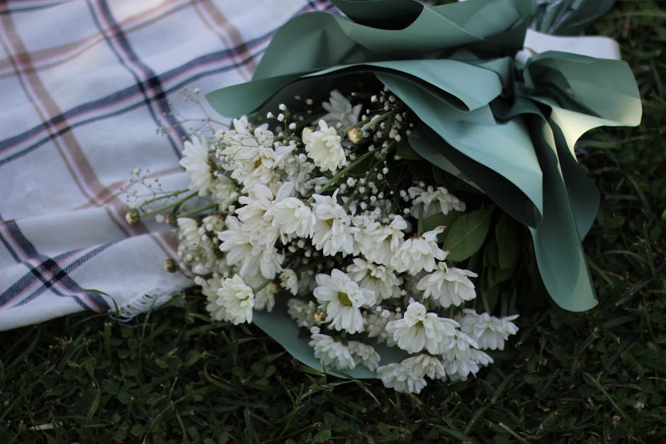 A bouquet of fresh white flowers, including daisies and small filler blooms, wrapped in elegant green paper with a satin ribbon, resting on a plaid cloth on the grass. The petals are soft and fully op