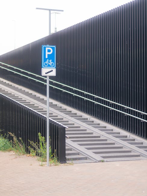 A flight of outdoor stairs with metal railings leading up alongside a tall, dark-colored vertical slat fence, adjacent to a paved pathway. Mounted on a pole at the base of the stairs is a blue parking