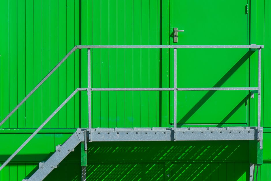 A close-up view of a metal staircase with grey railings leading up to a green wall with vertical panels. The stairs have perforated treads for grip, and the scene is illuminated by natural light, cast